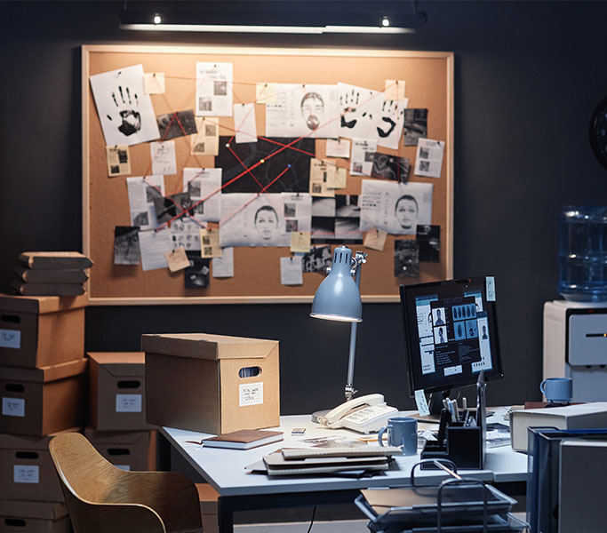 An office of a private investigator with a corkboard with various pictures on it, boxes of information, a desk, and a computer.